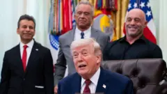 Donald Trump jókòó, ó tọwọ́ bọ inú ara wọn, àwọn ọkùnrin mẹta wà lẹ́yìn rẹ̀