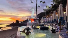 Pôr do sol com praia ao fundo e pessoas relaxando na piscina de um clube de praia em Bali