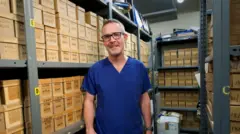 Um homem de uniforme cirúrgico azul e óculos está em pé em frente a prateleiras que vão do chão ao teto, repletas de caixas de papelão marrom