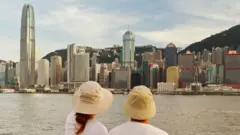 Eric e Emily aparecem de costas, usando chapéus claros do tipo bucket e camisetas brancas, enquanto observam o horizonte urbano à beira d’água