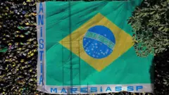 Foto feita de cima mostra uma grande bandeira do Brasil, nas margens está escrito "anistia" e "Maresias-SP, e dezenas de pessoas ao redor.