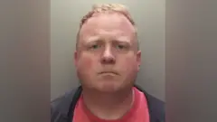 Robert Bush wit short, light brown hair show for dis straight‑on mugshot foto. Im no dey smile or frown, im wear dark jacket ontop red shirt. Di background dey plain and grey.