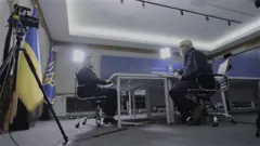 Zelensky está sentado à mesa com Jeremy Bowen. Ambos vestem camisas escuras. Eles estão em uma sala de conferências dentro do gabinete presidencial ucraniano, com bandeiras da Ucrânia atrás do presidente.