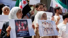 Önde iki kadın ailesinin fotoğraflarını turuyor, gerilerinde bir kadın ve birkaç adam daha var, Suriye bayrakları ve pankartlarla protesto ediyorlar. 