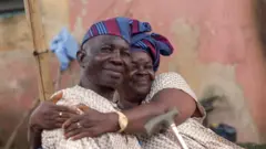 Amos ati Maria Adejinmi wọ leesi alawọ goolu iru kannaa, we gele ati fila aso oke alawọ buluu ati waini, wọn dijọ di mọ ara wọn, ti iyawo si n lo ọ̀pa rin.