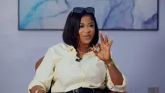 Imisioluwa Ayanwale to se ipo kinni nile Big Brother Naija fun ọdun 2025 joko lori aga kan, o na ọwọ osi soke bii ẹni to n ta ika, o fi irun wiigi dudu sori, o si fi igo akọ soke sori irun rẹ, o wọ asọ shaat alawọ miliki, o ọ sokoto alawọ buluu, to si lo bẹliti sori sokoto naa.