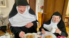 Duas freiras comem enquanto uma delas serve