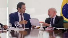 Fernando Haddad e Lula conversando e trocando papéis em uma mesa de trabalho, com a bandeira do Brasil ao fundo, no canto direito