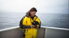 Lindy Weilgart, com casaco amarelo, em pé perto da proa de um barco, olhando para um binóculo