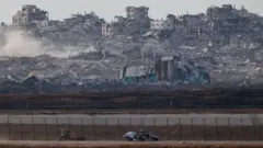 Um veículo militar israelense se move perto da fronteira entre Israel e Gaza. Ao fundo, há uma massa de destroços e fumaça de prédios destruídos em Gaza