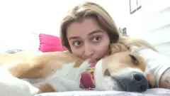 Catalina apoia a cabeça sobre um cachorro que dorme na cama e olha para a câmera