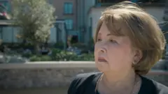 krupni kada starije žene, Japanke, koja gleda zamišljeno u stranu, iza nje mutna pozadina, ulica, drvo, kuće.