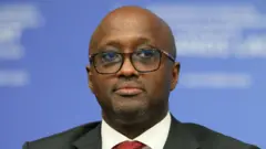 Minisitiri w'ububanyi n'amahanga w'u Rwanda Olivier Nduhungirehe yambaye ikositimu y'umukara, ishati y'umweru na karuvati y'umutuku irimo utubara tw'umukara. Arareba muri 'camera' asa nk'ureba hejuru gato. Yari yitabiriye inama ku mahoro n'uburumbuke mu karere k'ibiyaga bigari, yabereye i Paris mu Bufaransa, ku itariki ya 30 Ukwakira (10) mu 2025.