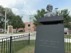 Busto del general José Artigas, prócer de Uruguay, en la plaza frente al ayuntamiento de Elizabeth. A la izquierda se ve un mástil con una bandera de Estados Unidos y, de fondo, la fachada de una escuela secundaria.