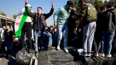 مجموعة من المتظاهرين الشباب يتظاهرون أمام الكاميرات وهم يدوسون على وجه تمثال يصور أحد أقارب الرئيس السوري المخلوع