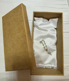 Foto de uma caixa de madeira menor que uma caixa de sapatos, aberta, contendo um pequeno terço sobre uma almofada branca