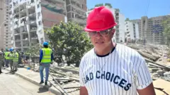 Ein Mann mit rotem Projekthelm und Sonnenbrille stand auf der Straße vor den Trümmern eines eingestürzten Gebäudes, in der Nähe mehrere Beamte in Warnwesten.