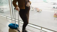 Mujer en embarazo espera en un pasillo de un aeropuerto mientras mira los aviones en la pista. Viste elegantemente y lleva una maleta beige. 