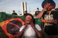 Dois indígenas protestando ao lado de bandeira do Brasil com aparência ensanguentada e prédios do Congresso Nacional atrás