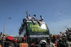 Des joueurs de l'équipe nationale du Sénégal sur le toit d'un car sur lequel est écrit "Champions d'Afrique 2025, senecartours" lors de la parade avec le trophée d ela CAN dans les rues de Dakar. Une foule de supporteurs et de policiers autour du véhicule.