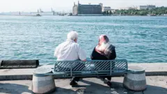 Kadıköy'de Haydarpaşa Garı'nı gören bir açıdan deniz kenarındaki banka oturmuş yaşlı bir çift. Arkadan gördüğümüz çiftten soldaki erkek olanı üzerinde kısa kollu bir gömlekle otururken sağdaki kadının üstünde ise siyah bir kıyafet ve desenli bir başörtüsü var