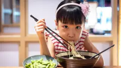 Menina oriental comendo macarrão com palitos e vegetais