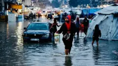 د بازار یوه څنډه چې پر سړک اوبه ډنډ دي، موټر او خلک پکې روان دي او تر څنګ یې کېږدۍ ښکاري چې اوبه ورننوتلې دي.