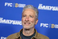 Jon Stewart, con chaqueta vaquera en tonos camel una camiseta negra con un fondo de color azul con letras donde se promociona su programa "The Daily Show".