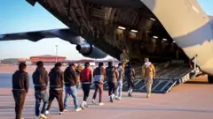 Indege ya gisirikare ya Leta Zunze Ubumwe za Amerika iri ku kibuga cy’indege, n’abakozi bo ku kibuga cy'indege n'abantu bagiye kuyinjiramo ngo bajyanwe mu kindi gihugu