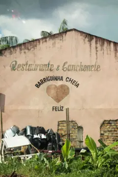 Fachada lateral com uma pintura feita à mão, onde se lê "Restaurante & Lanchonete Barriguinha Cheia Feliz", com o desenho de um coração. Embaixo da pintura na fachada, se vê uma pilha de entulho e mato crescido