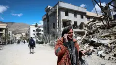 がれきの山と化した破壊された建物の前、携帯電話を手にして話す女性。晴天の下、遠方には小高い丘が見える