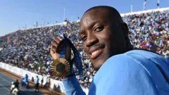 Letsile Tebogo regarde en arrière, brandit sa médaille d'or olympique de Paris 2024 et sourit à la caméra, tandis qu'une tribune remplie de milliers de supporters est visible au loin derrière lui.