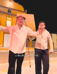 Dois homens vestidos com camisa branca e calça preta. Um deles usa um chapéu típico do Nordeste