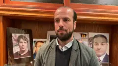 المحامي فادي العبار، ممثل رابطة عائلات قيصر في دمشق تظهر خلفه مجموعة صور.