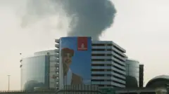 Smoke rising from an area near Dubai International Airport is seen through a vehicle’s windshield after a drone attack hit a fuel tank. In the foreground, an Emirates poster portraying a female flight attendant covers almost the entire façade of a building