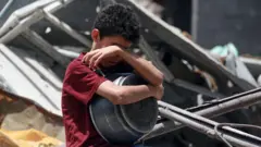 Um menino cobre o rosto segurando uma panela vazia enquanto tenta coletar ajuda alimentar em Gaza no início de maio