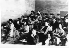 Un grupo de estudiantes, entre ellos hay varias mujeres, en un salón de clases. Todos están sentados en pupitres de madera