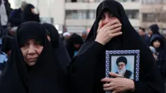 Two women in black at a rally in Tehran. One holding a portrait of the assassinated Ayatollah Ali Khamenei and crying