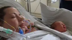 A woman in a hospital bed with the white sheet pulled up to her chin. Her hair, which is brown, is pulled back from her face. She is not conscious and there are tubes entered into her nostrils, and by her mouth. A newborn baby, with a small amount of dark hair and wearing a white babygrow, is lying next to her with a pillow along side and with a white blanket around it. A person is visible standing to the right of the frame, wearing dark blue, but only part of their mid-section and hands can be seen.