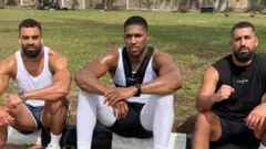 Anthony Joshua siddon between Sina Ghami and Latif Ayodele as all wear training kit, wit one grassy area behind di three men.