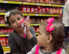 Duas meninas pequenas sentam em um carrinho de compras; uma delas segura vários tipos de doces. As duas usam laços no cabelo. Atrás delas, as prateleiras do supermercado estão cheias de guloseimas.