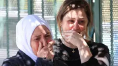 Beyaz başörtülü bir kadınla sarışın bir kadın bir noktaya bakarak ağlıyor.