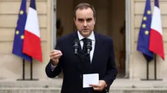 Sébastien Lecornu gestures as he speaks into a microphone outside of a building, with French and EU flags either side of him.