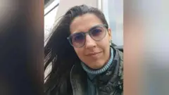 Vitória está olhando diretamente para a câmera. Ela veste uma blusa de gola alta azul, um casaco cáqui ou escuro, usa óculos e tem cabelos castanhos.