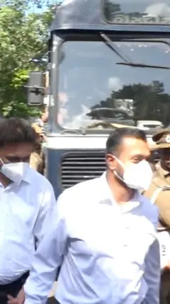 නිල් පාට බස් රථයක් ඉදිරියෙන් මුඛ ආවරණ දමාගත් පිරිමි දෙදෙනෙකු එන අයුරු පෙනේ.