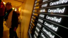 Vista de um terminal de aeroporto com um grande painel eletrônico de partidas com várias listas de voos, todos marcados como “Cancelled” (cancelado) em letras brancas sobre fundo escuro, com números de voos visíveis