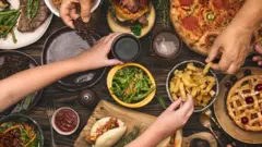 Mãos de diversas pessoas pegam alimentos, como batatas fritas, pizza, saladas e carne, de tigelas sobre uma mesa de madeira