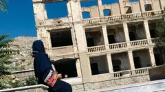 منظر جانبي لنادية بالحجاب الأسود أمام مبنى متضرر
