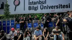 Bilgi Üniversitesi'nin girişinde oturan bir grup öğrencinin arkasında polisler kalkanlarıyla ayakta duruyor