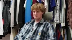 أليكسي هامبلين يجلس على كرسي وخلفه مجموعة من الملابس